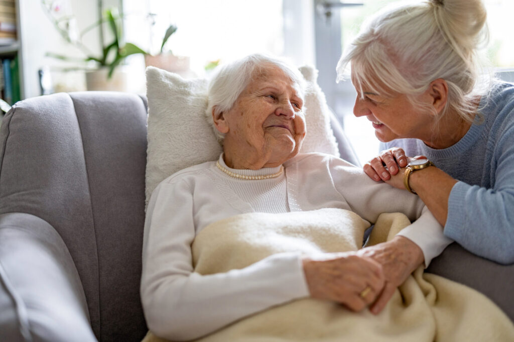 Older woman lounging in a comfortable chair. Her daughter is kneeling beside her. They are smiling at one another.